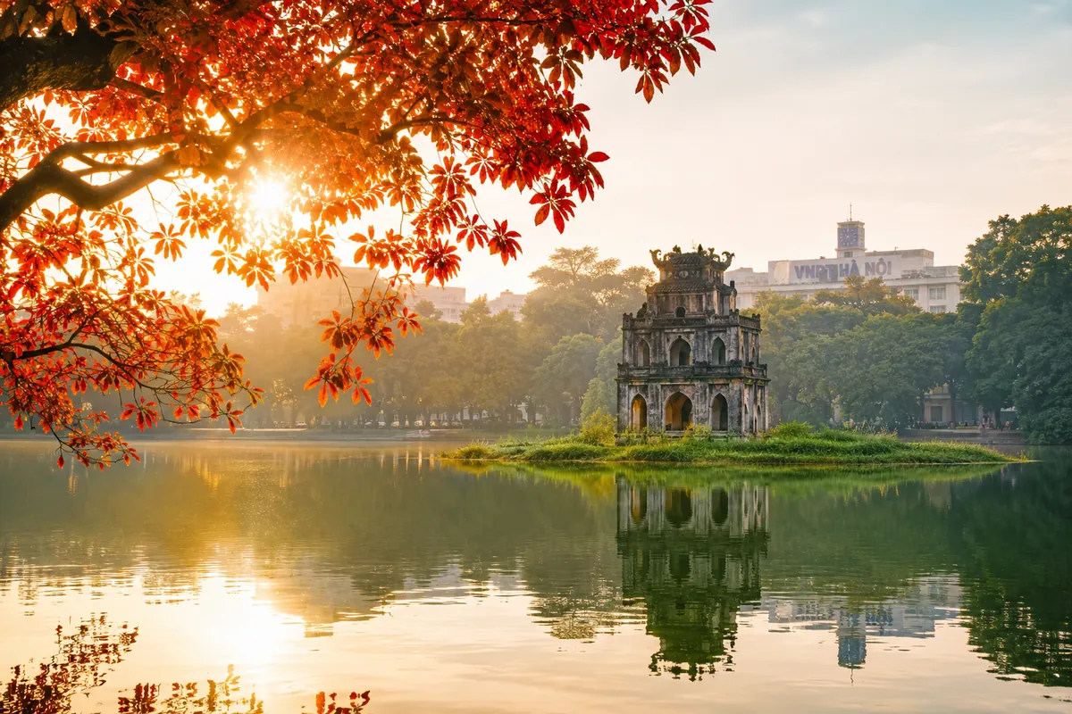 The Turtle Tower on Hoan Kiem Lake in Hanoi at sunrise, framed by red leaves with the Old Quarter behind