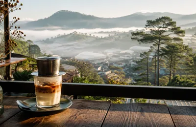 Vietnamese phin coffee dripping into a glass on a wooden table overlooking misty Da Lat hills